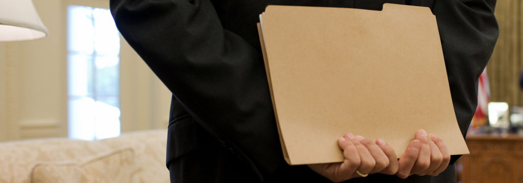 A man in a dark suit holds a manilla file folder behind his back
